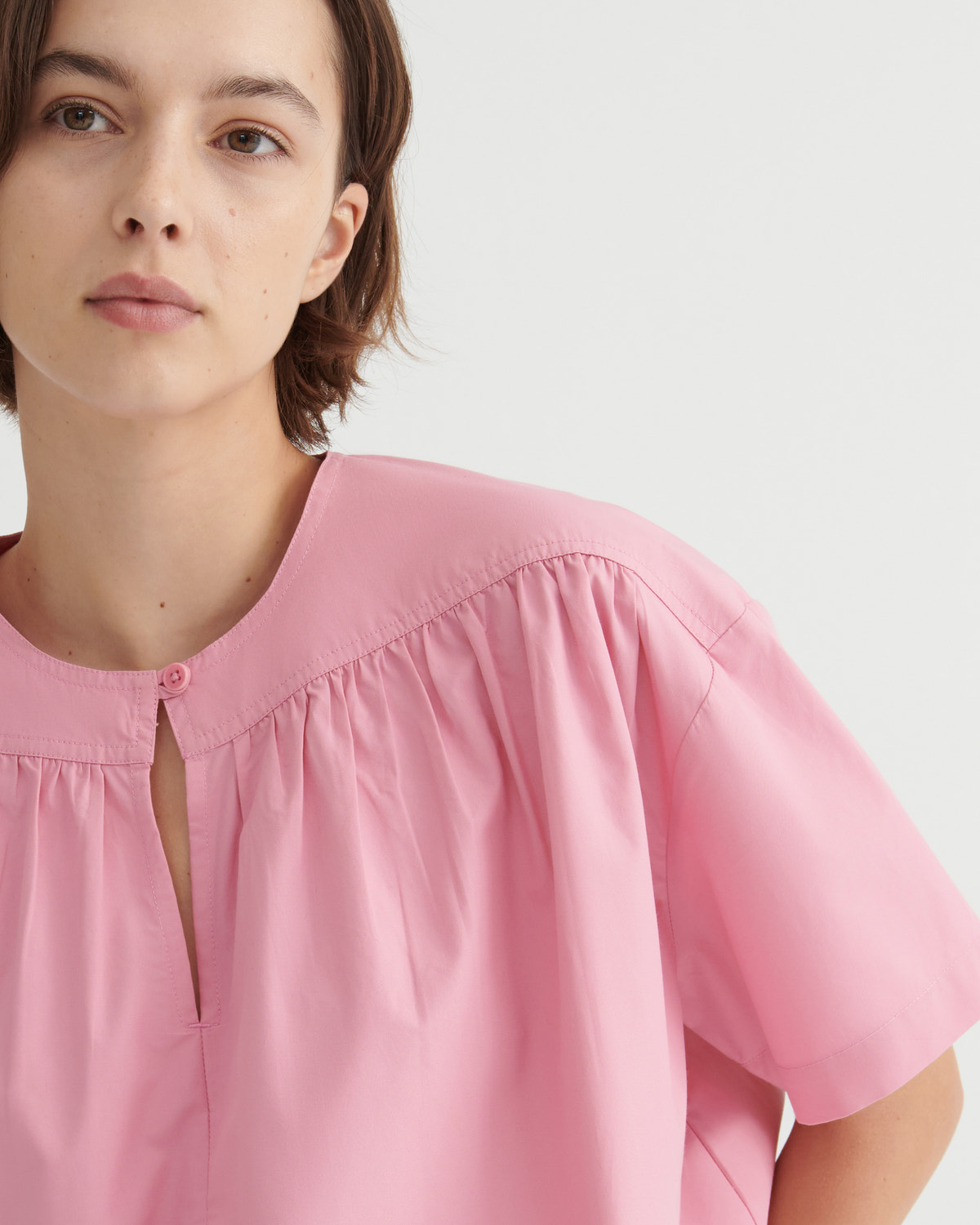 A close-up of a model wearing a light pink short-sleeve blouse with a gathered round neckline and a small front keyhole cutout.