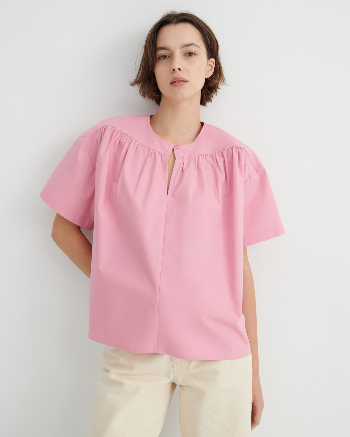 A woman wearing a light pink short-sleeve blouse with a gathered round neckline and a small front slit, paired with cream pants, standing against a plain light gray wall.
