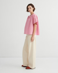 A model stands in profile wearing a light pink short-sleeve blouse with a gathered neckline, paired with wide-leg cream trousers and red sandals, against a clean white studio background.