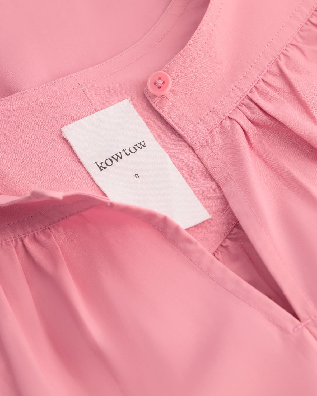 Close-up of a pink garment with a visible white Kowtow brand label and a matching pink button at the neckline.