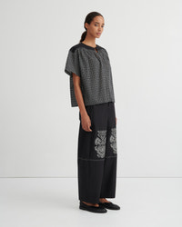 A model stands in a studio wearing a black and white micro-check short-sleeve top paired with black wide-leg trousers featuring white embroidered details on the lower legs, and black flat shoes.