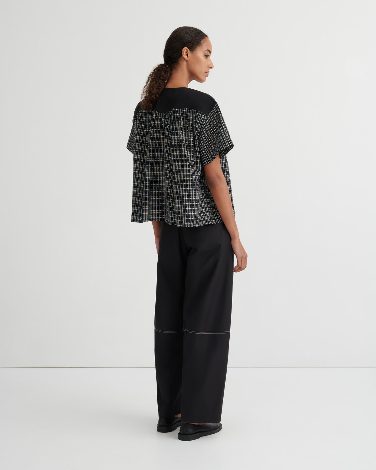 A model is shown from the back wearing a black and white checkered short-sleeve top with a solid black yoke, paired with black wide-leg trousers, against a plain white studio background.