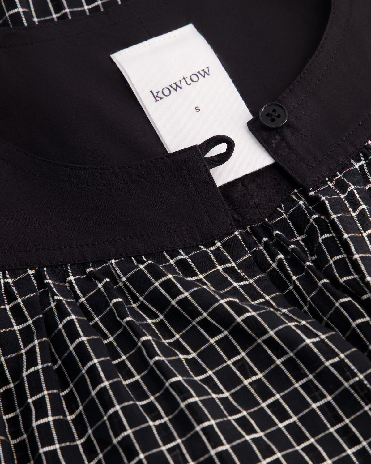 Close-up of a Kowtow garment showing a black collar with a white brand tag, a black button, and a black-and-white grid patterned fabric below.