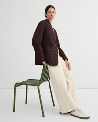 A model wearing a dark brown blazer with contrast topstitching, cream wide-leg trousers, and white sneakers, leaning against a green metal chair in a studio setting.