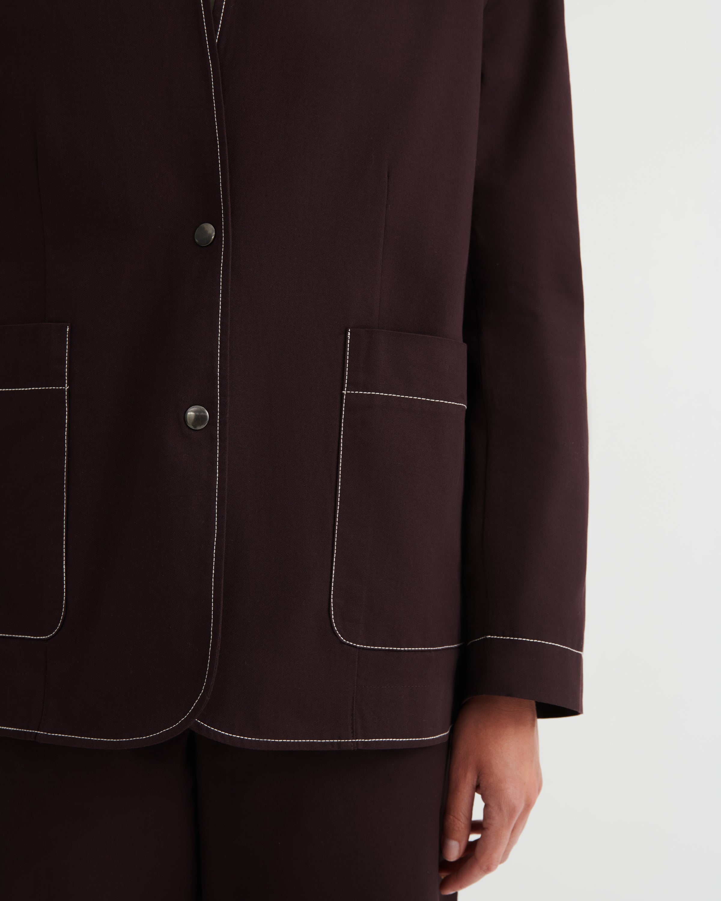 Close-up of a dark brown tailored jacket with white contrast stitching, featuring two large front patch pockets and a button closure, worn by a model against a light gray background.