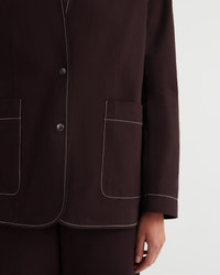 Close-up of a dark brown tailored jacket with white contrast stitching, featuring two large front patch pockets and a button closure, worn by a model against a light gray background.