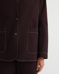 Close-up of a dark brown tailored jacket with white contrast stitching, featuring two large front patch pockets and a button closure, worn by a model against a light gray background.