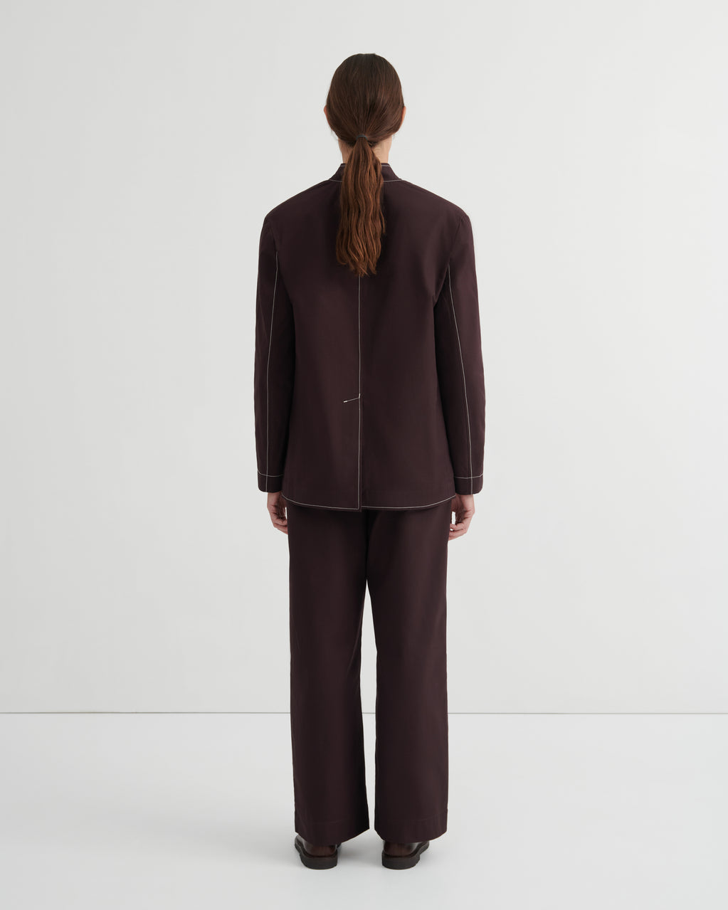 A model viewed from the back wearing a dark brown tailored jacket and matching wide-leg trousers against a plain light gray studio background.