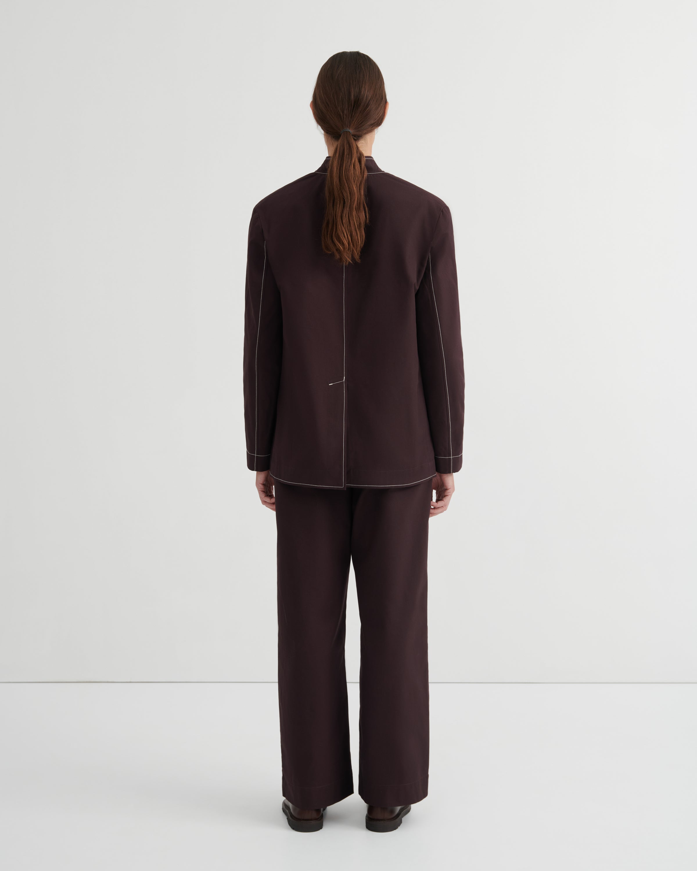 A model viewed from the back wearing a dark brown tailored jacket and matching wide-leg trousers against a plain light gray studio background.