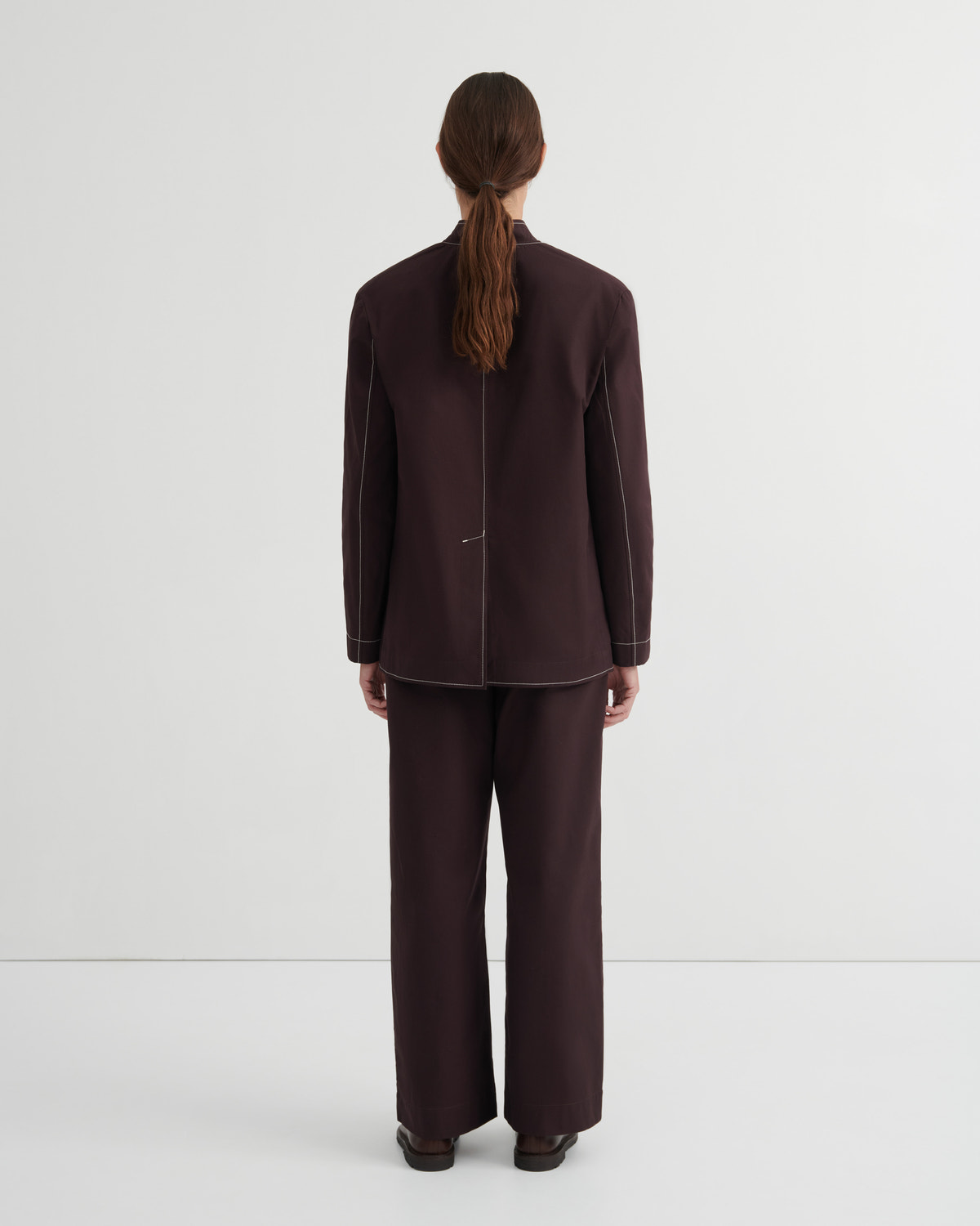 A model viewed from the back wearing a dark brown tailored jacket and matching wide-leg trousers against a plain light gray studio background.