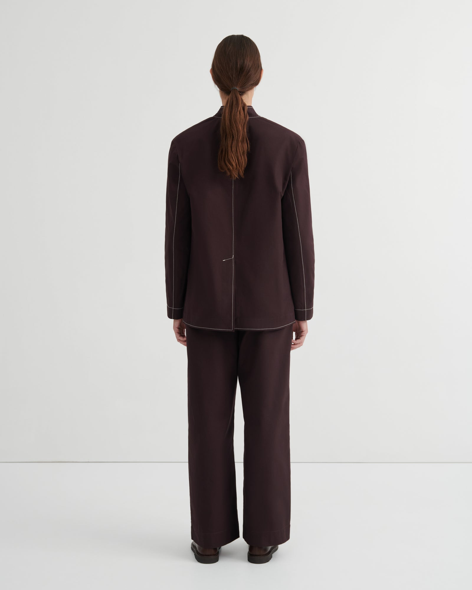 A model viewed from the back wearing a dark brown tailored jacket and matching wide-leg trousers against a plain light gray studio background.