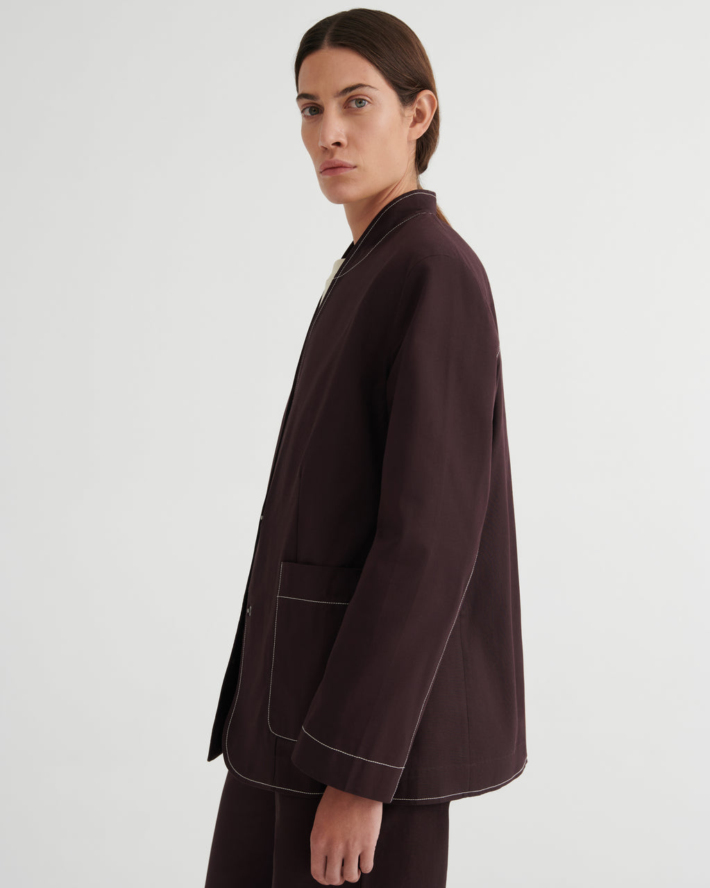 A model shown from the side in a dark brown blazer with contrasting light topstitching, paired with matching dark trousers, against a plain light gray studio background.