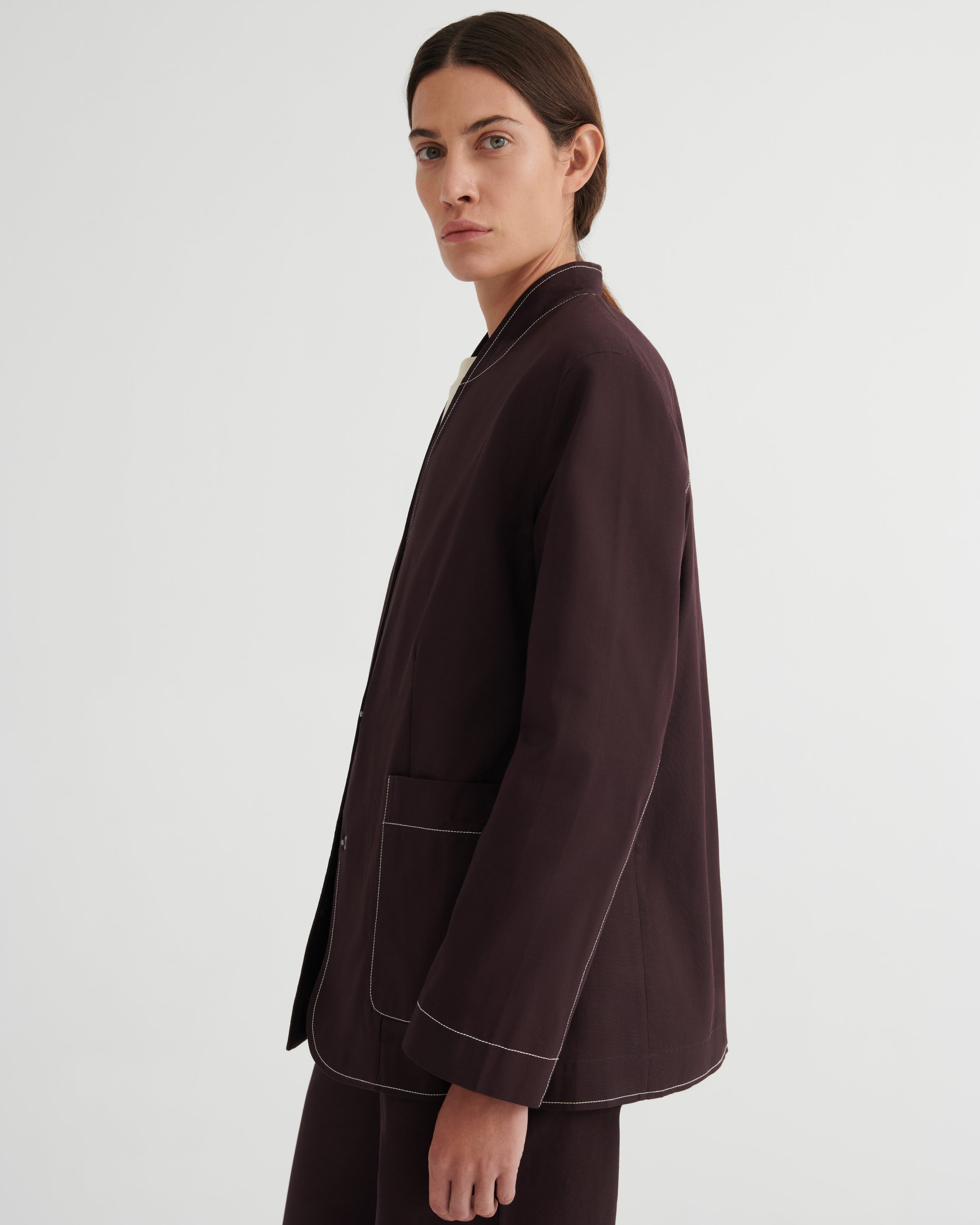 A model shown from the side in a dark brown blazer with contrasting light topstitching, paired with matching dark trousers, against a plain light gray studio background.