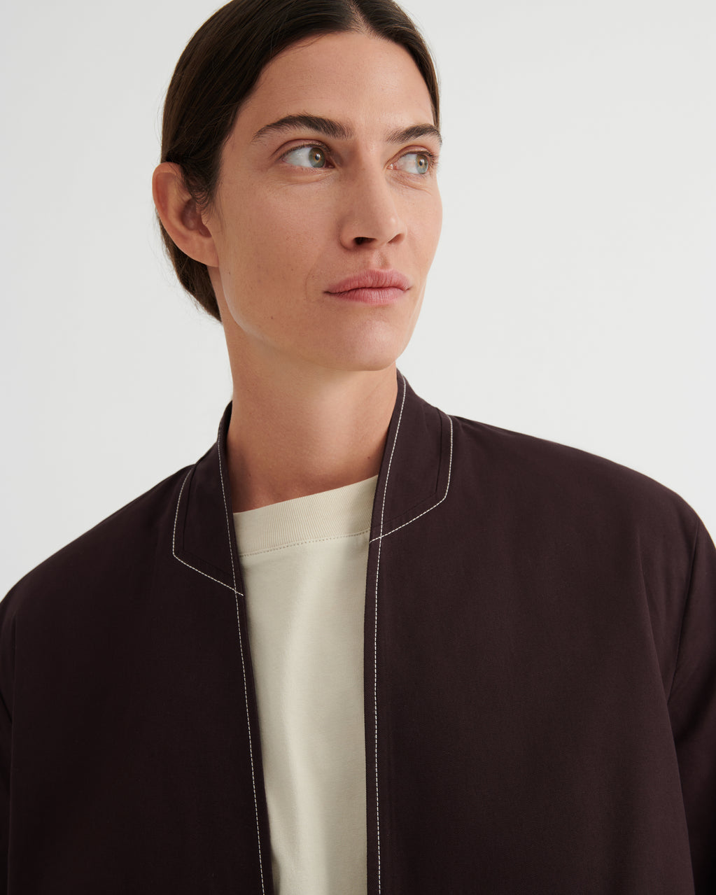A close-up of a model wearing a dark brown blazer with white contrast stitching over a cream crewneck shirt, set against a plain light gray background.