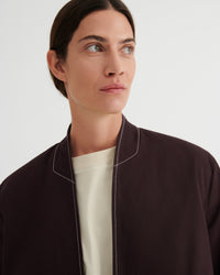 A close-up of a model wearing a dark brown blazer with white contrast stitching over a cream crewneck shirt, set against a plain light gray background.