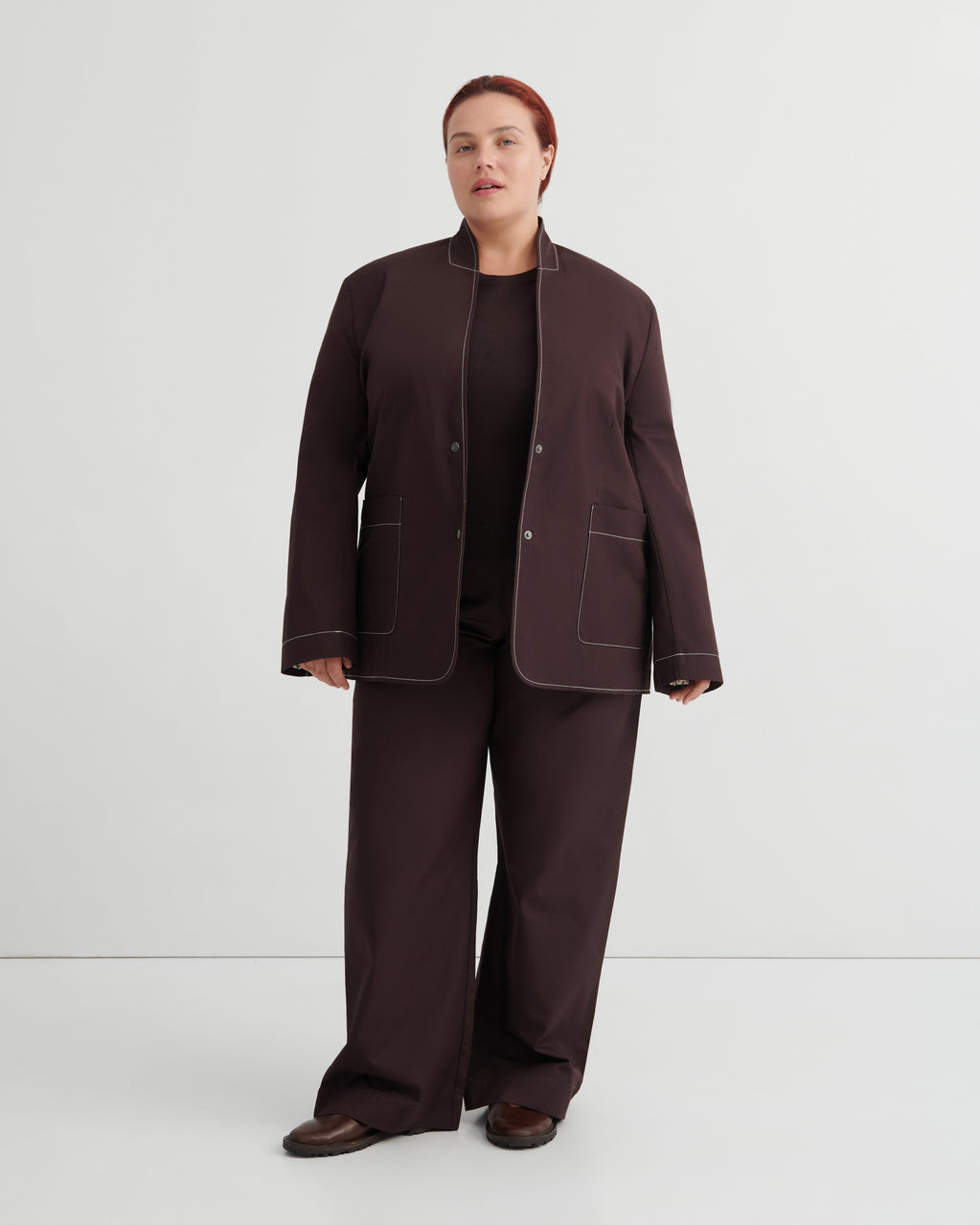 A model stands against a light gray studio background wearing a dark brown matching set consisting of a collarless blazer with contrast topstitching, wide-leg trousers, and a black top.