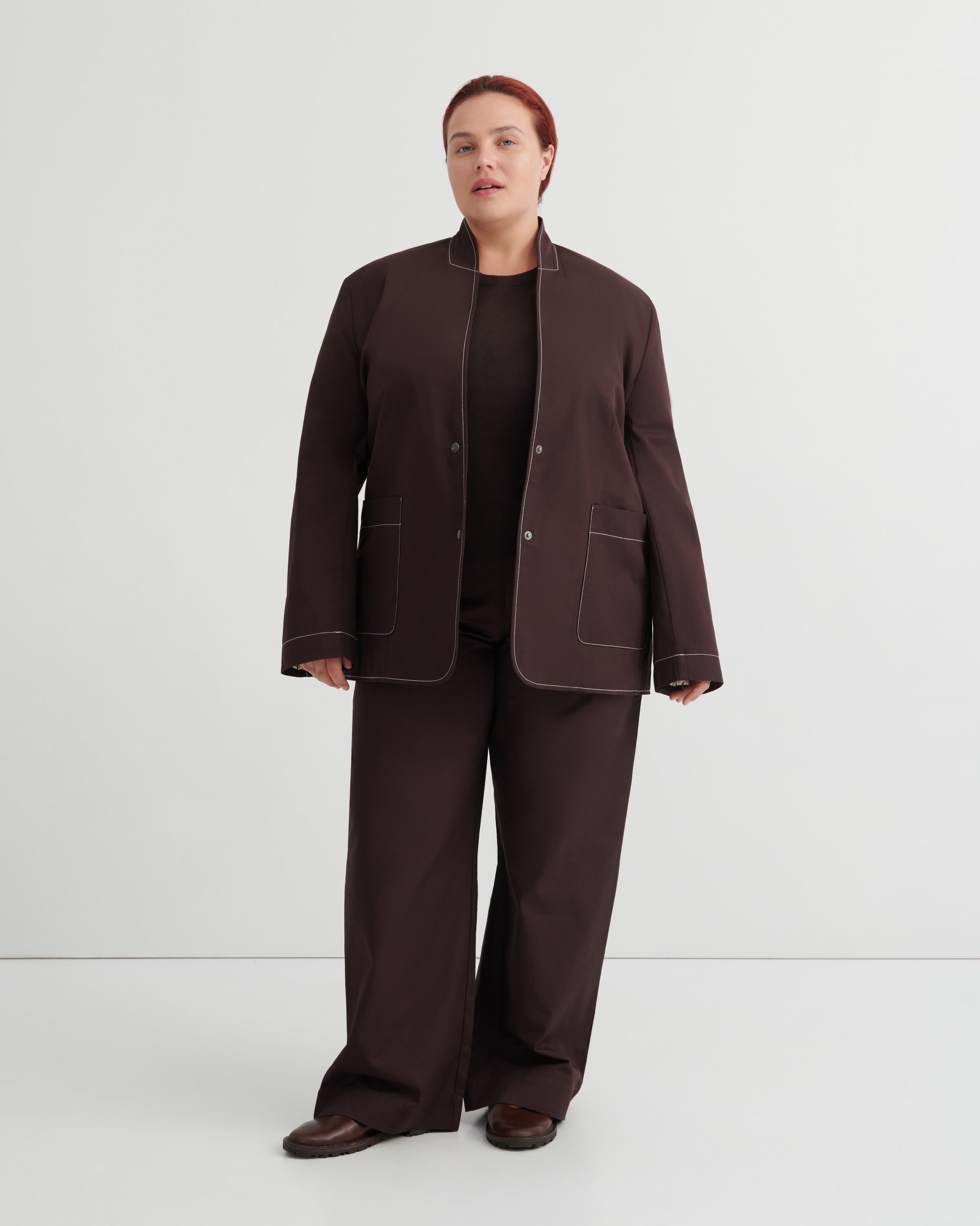 A model stands against a light gray studio background wearing a dark brown matching set consisting of a collarless blazer with contrast topstitching, wide-leg trousers, and a black top.