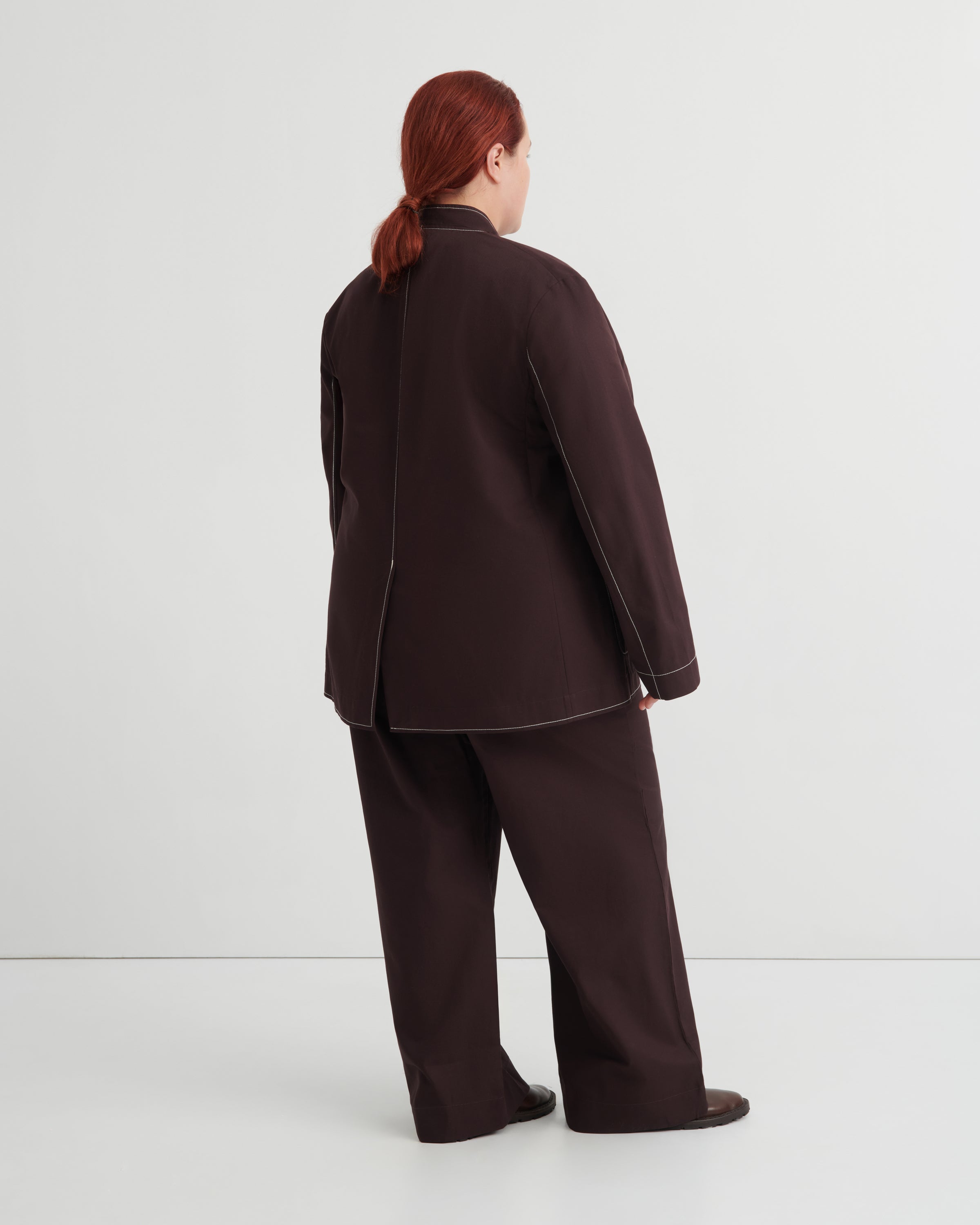 A model with red hair tied back is shown from the back wearing a dark brown two-piece suit with a mandarin collar jacket and wide-leg trousers, set against a plain light gray studio background.