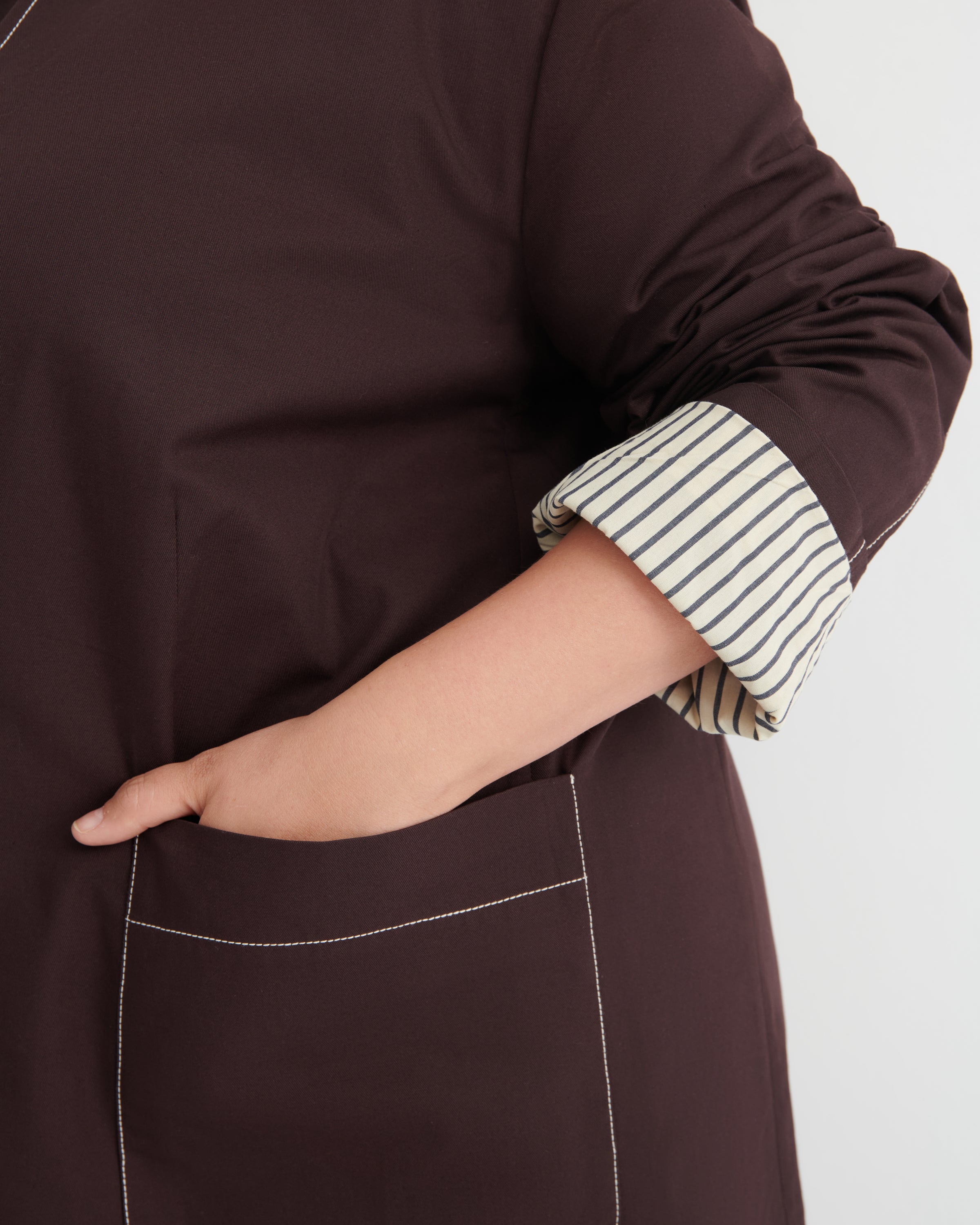 A close-up of a person wearing a dark brown blazer with a striped lining rolled up at the sleeve, showing a hand in a pocket against a light gray background.