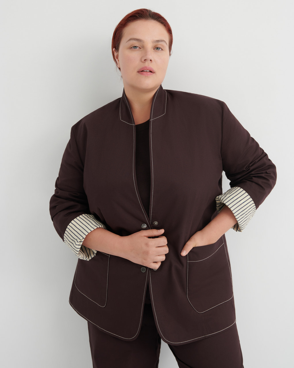 A woman modeling a dark brown blazer with contrasting topstitching and striped cuffs, paired with matching dark brown trousers against a light gray studio background.