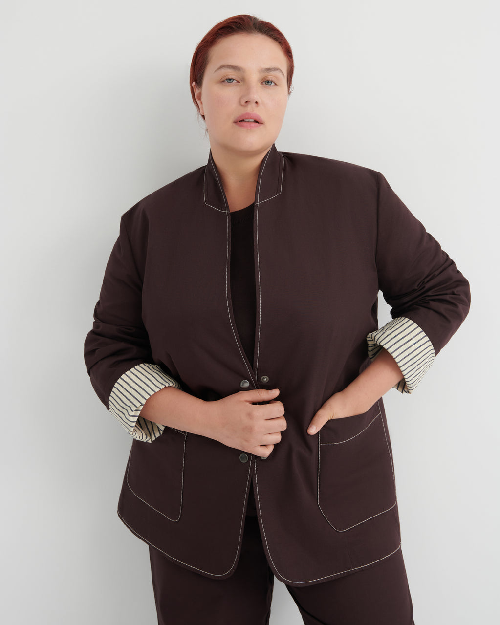 A woman modeling a dark brown blazer with contrasting topstitching and striped cuffs, paired with matching dark brown trousers against a light gray studio background.
