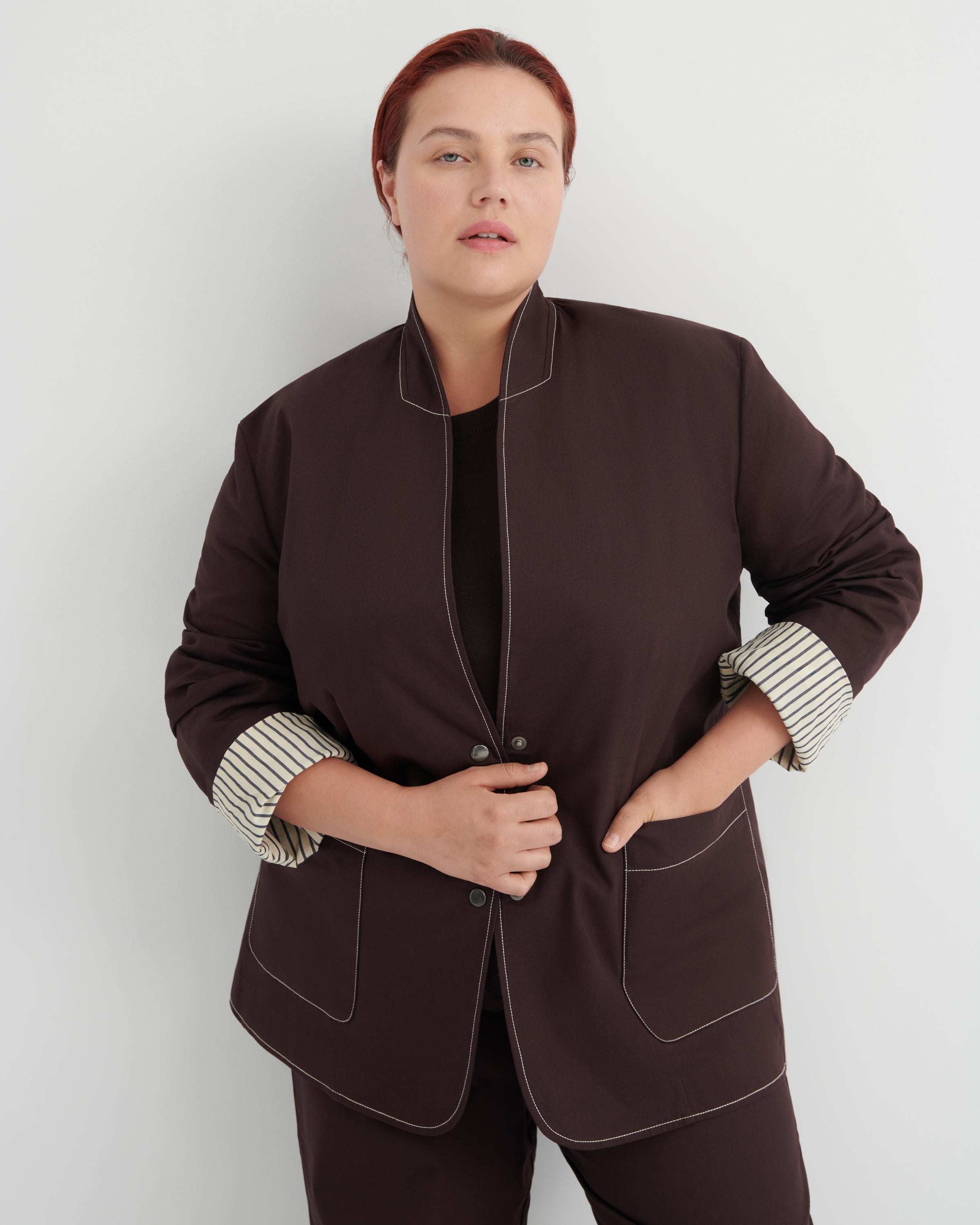 A woman modeling a dark brown blazer with contrasting topstitching and striped cuffs, paired with matching dark brown trousers against a light gray studio background.