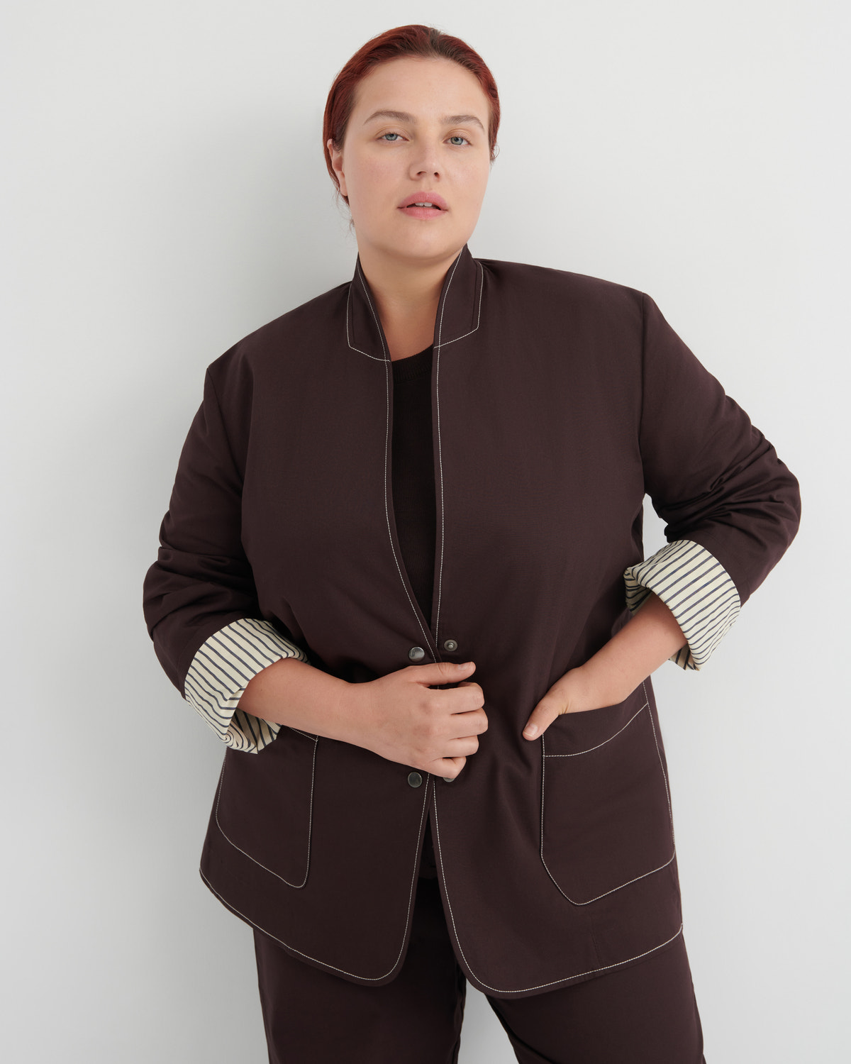 A woman modeling a dark brown blazer with contrasting topstitching and striped cuffs, paired with matching dark brown trousers against a light gray studio background.