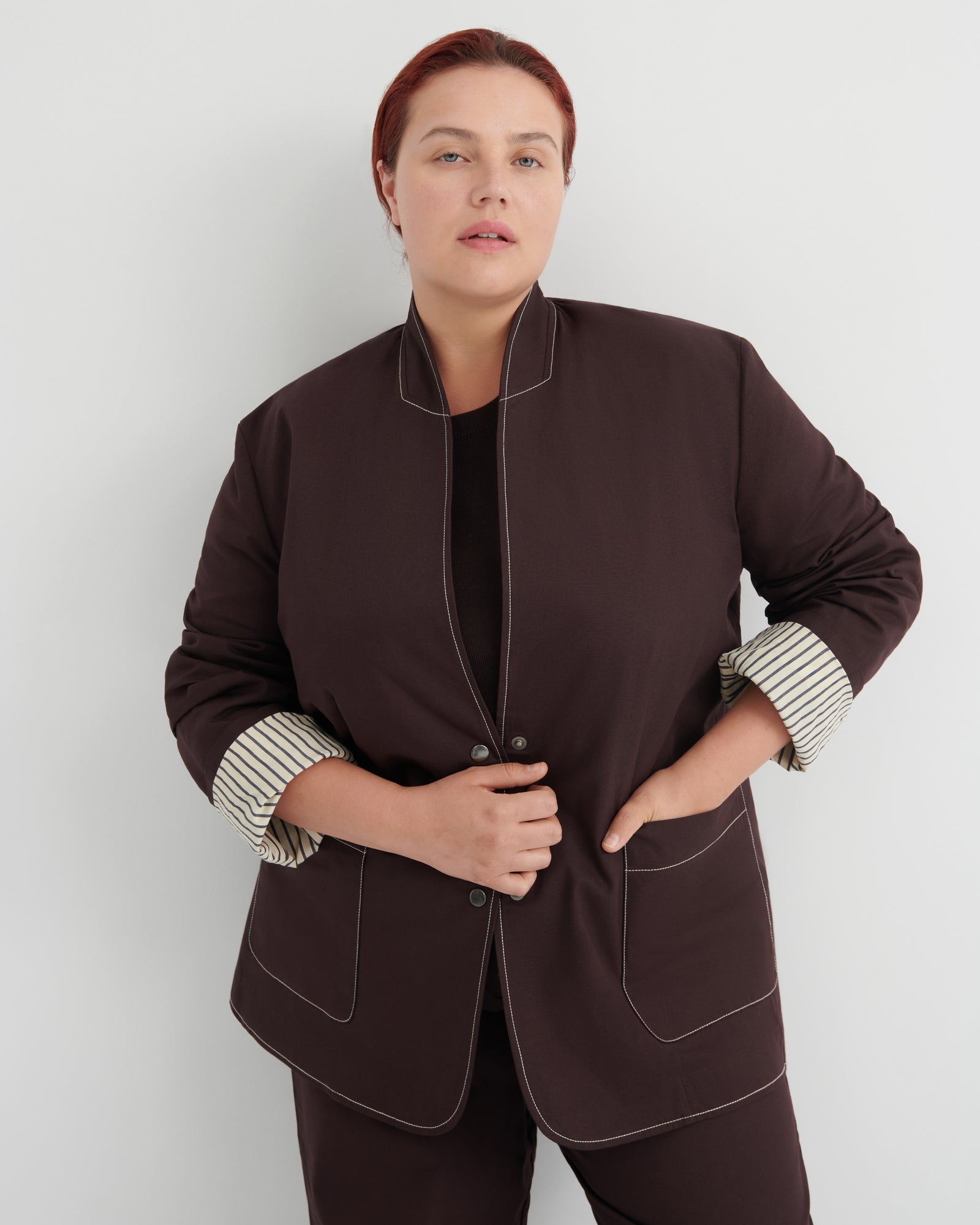 A woman modeling a dark brown blazer with contrasting topstitching and striped cuffs, paired with matching dark brown trousers against a light gray studio background.