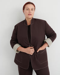 A woman modeling a dark brown blazer with contrasting topstitching and striped cuffs, paired with matching dark brown trousers against a light gray studio background.