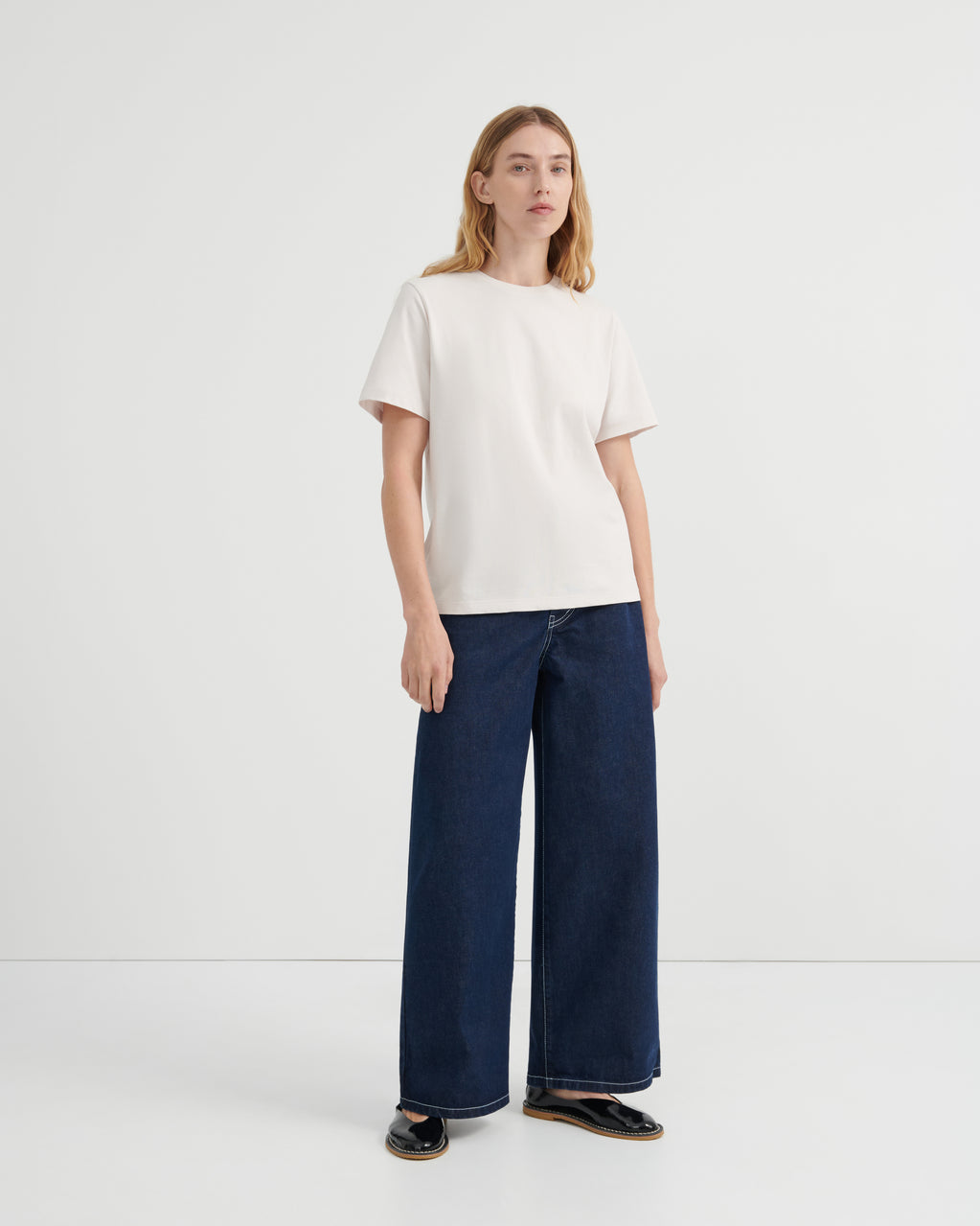A woman wearing an off-white short-sleeve crewneck t-shirt and dark blue wide-leg jeans.