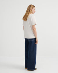 A woman with long blonde hair is shown from the back wearing a off-white short-sleeve t-shirt and dark blue wide-leg jeans.