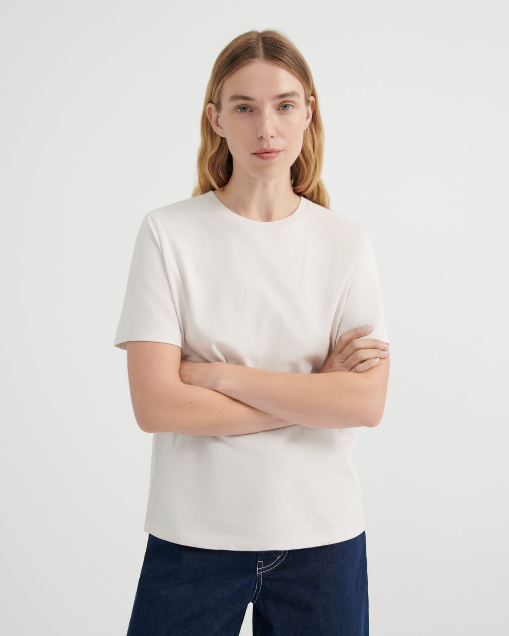 A woman with blonde hair wearing a plain off-white short-sleeve crewneck t-shirt and dark blue jeans with her arms crossed.