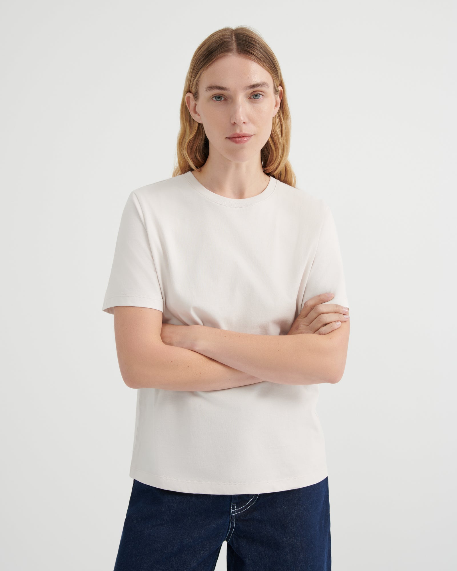 A woman with blonde hair wearing a plain off-white short-sleeve crewneck t-shirt and dark blue jeans with her arms crossed.