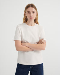 A woman with blonde hair wearing a plain off-white short-sleeve crewneck t-shirt and dark blue jeans with her arms crossed.