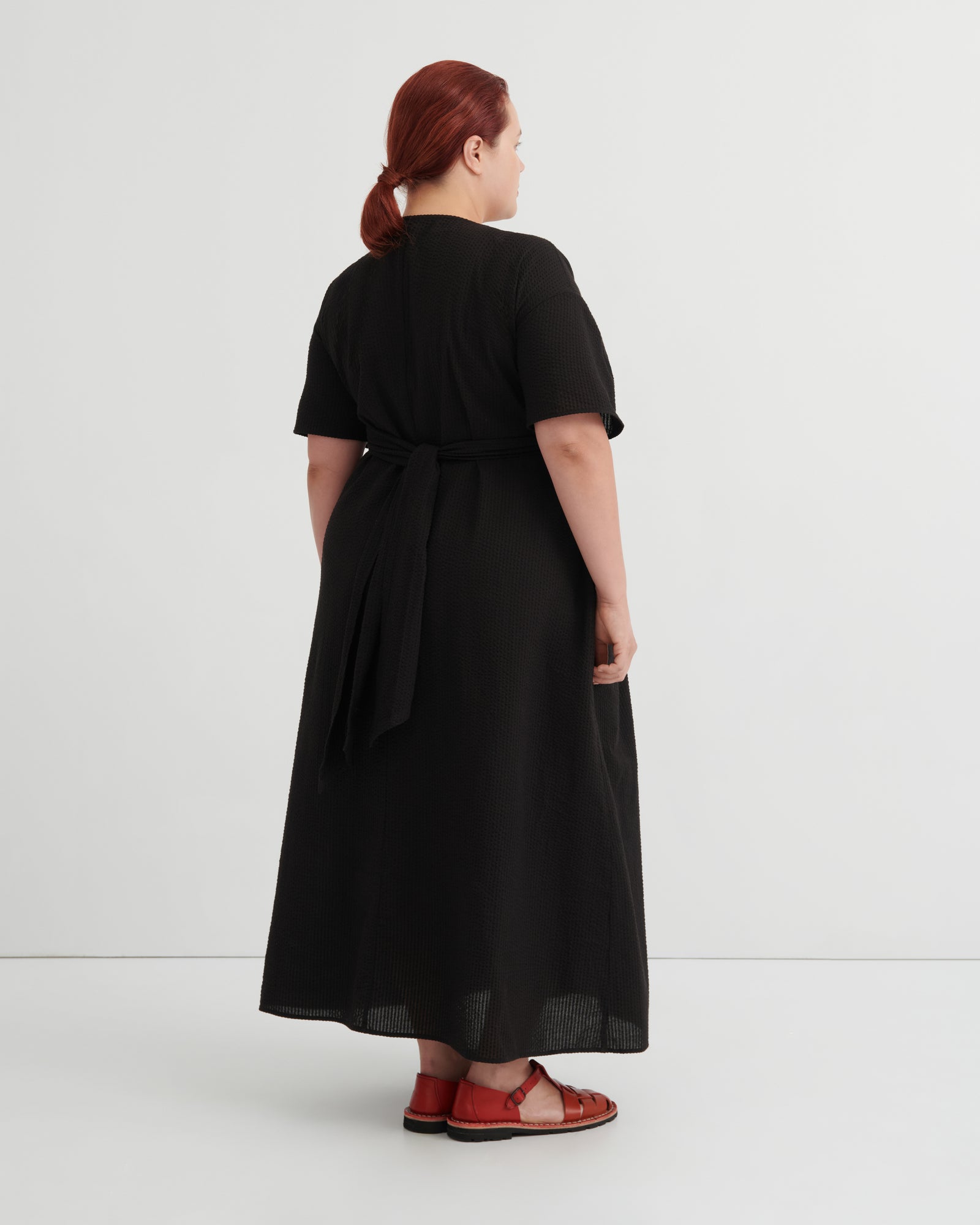 A woman with red hair tied back is shown from the back wearing a black short-sleeve midi dress with a tie waist, paired with red sandals.