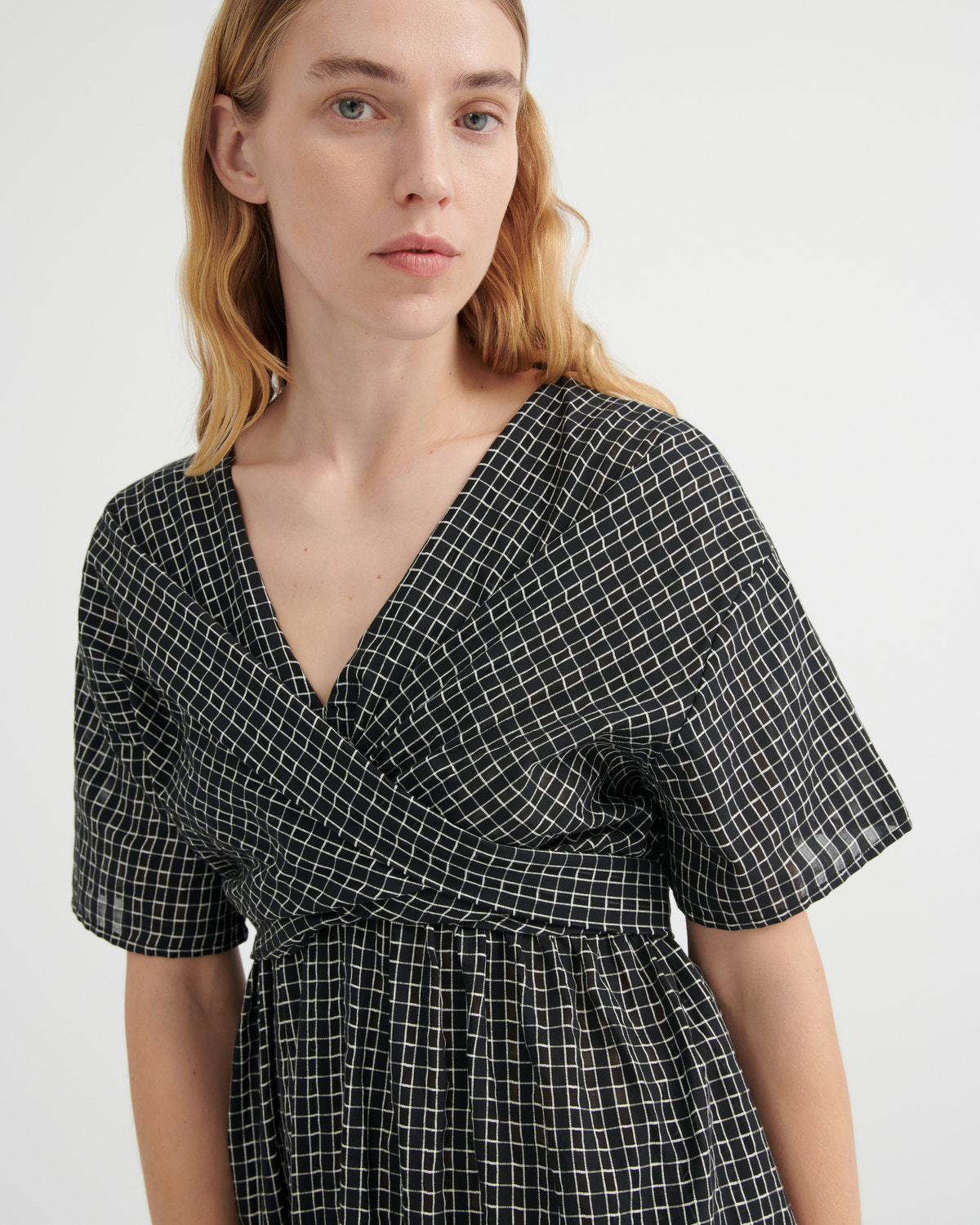 A woman wearing a black and white grid-patterned wrap dress with short flutter sleeves and a V-neckline, photographed against a clean white studio background.