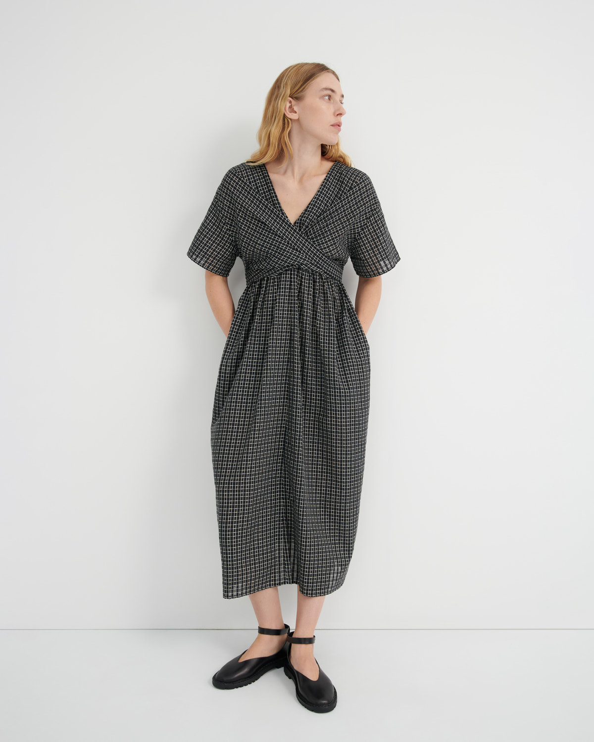 A woman modeling a short-sleeve black and white checkered midi dress with a wrap-style V-neck and side pockets, paired with black Mary Jane shoes, against a plain white studio background.