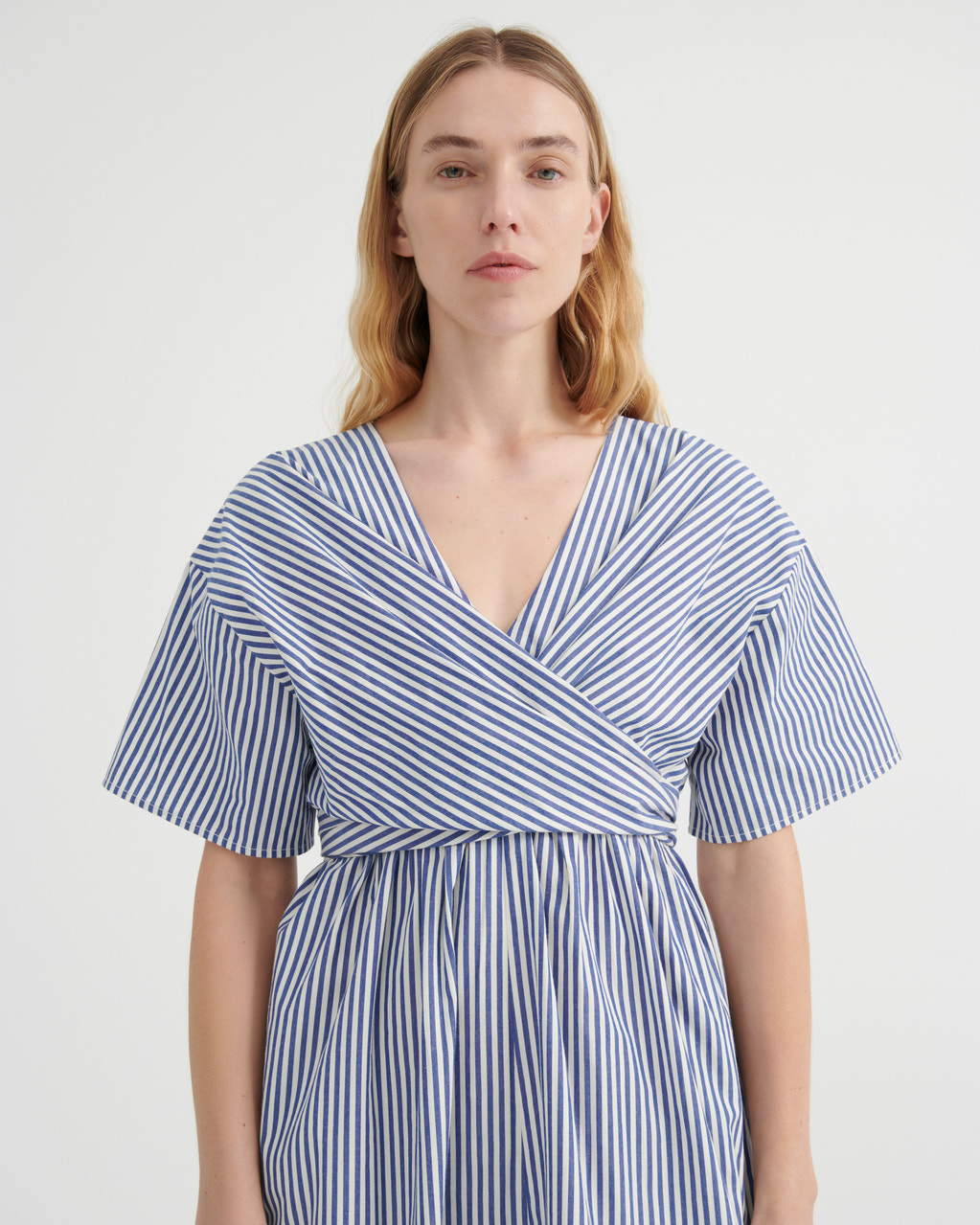 A woman modeling a blue and white vertically striped wrap dress with short flutter sleeves and a V-neckline, photographed against a plain light gray studio background.