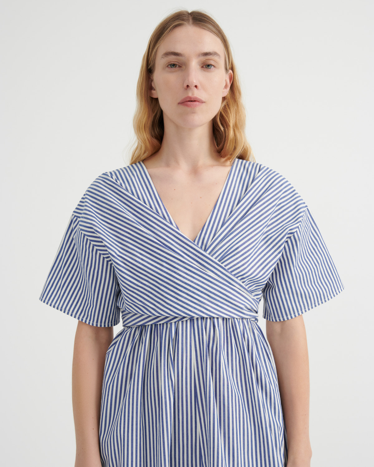 A woman modeling a blue and white vertically striped wrap dress with short flutter sleeves and a V-neckline, photographed against a plain light gray studio background.