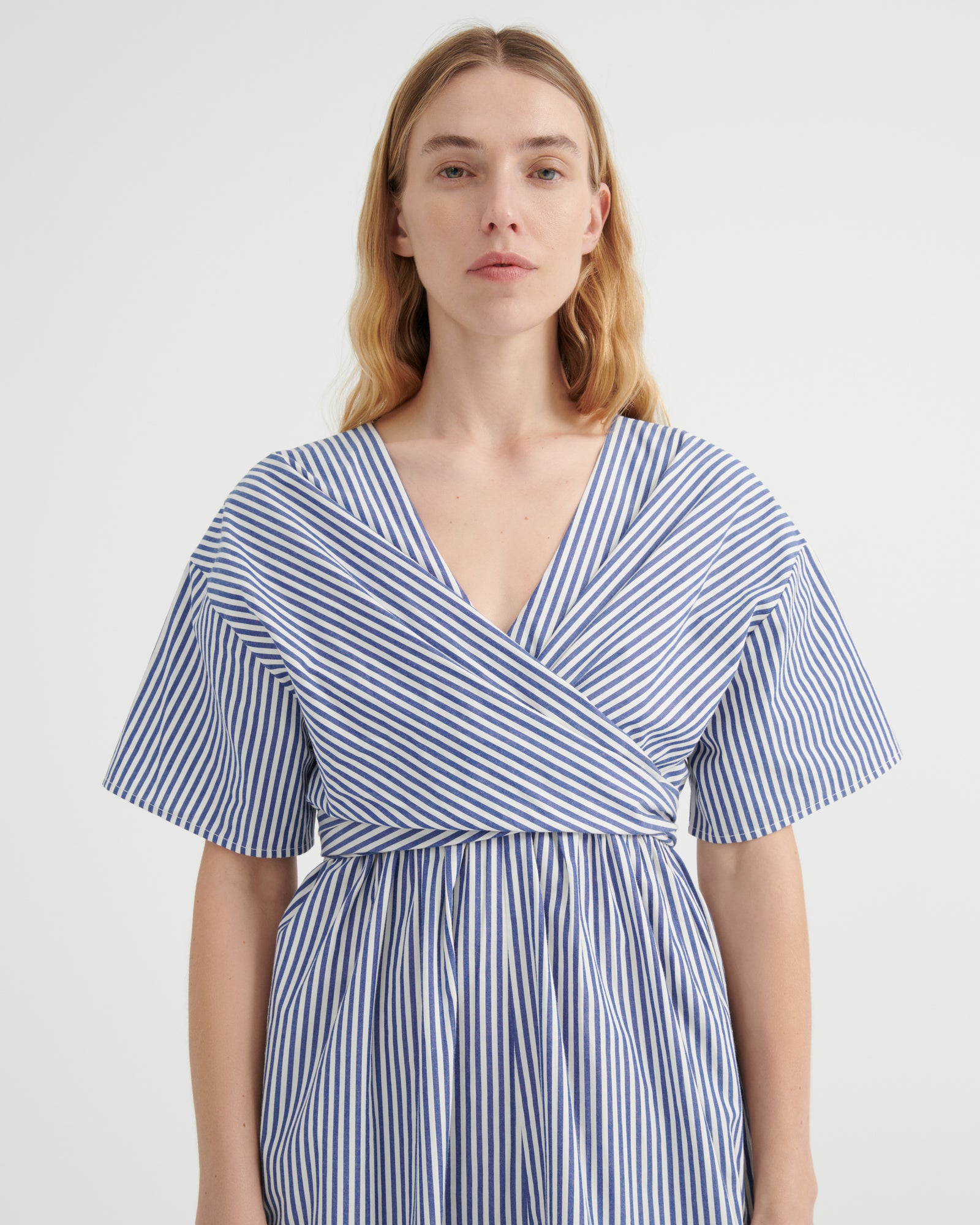 A woman modeling a blue and white vertically striped wrap dress with short flutter sleeves and a V-neckline, photographed against a plain light gray studio background.