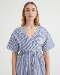 A woman modeling a blue and white vertically striped wrap dress with short flutter sleeves and a V-neckline, photographed against a plain light gray studio background.
