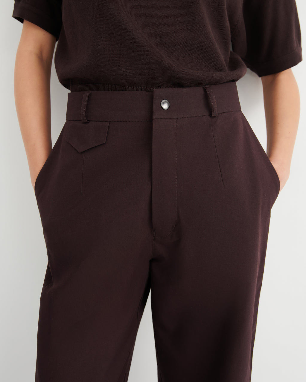 A close-up of a person wearing dark brown tailored trousers and a dark brown short-sleeve top.