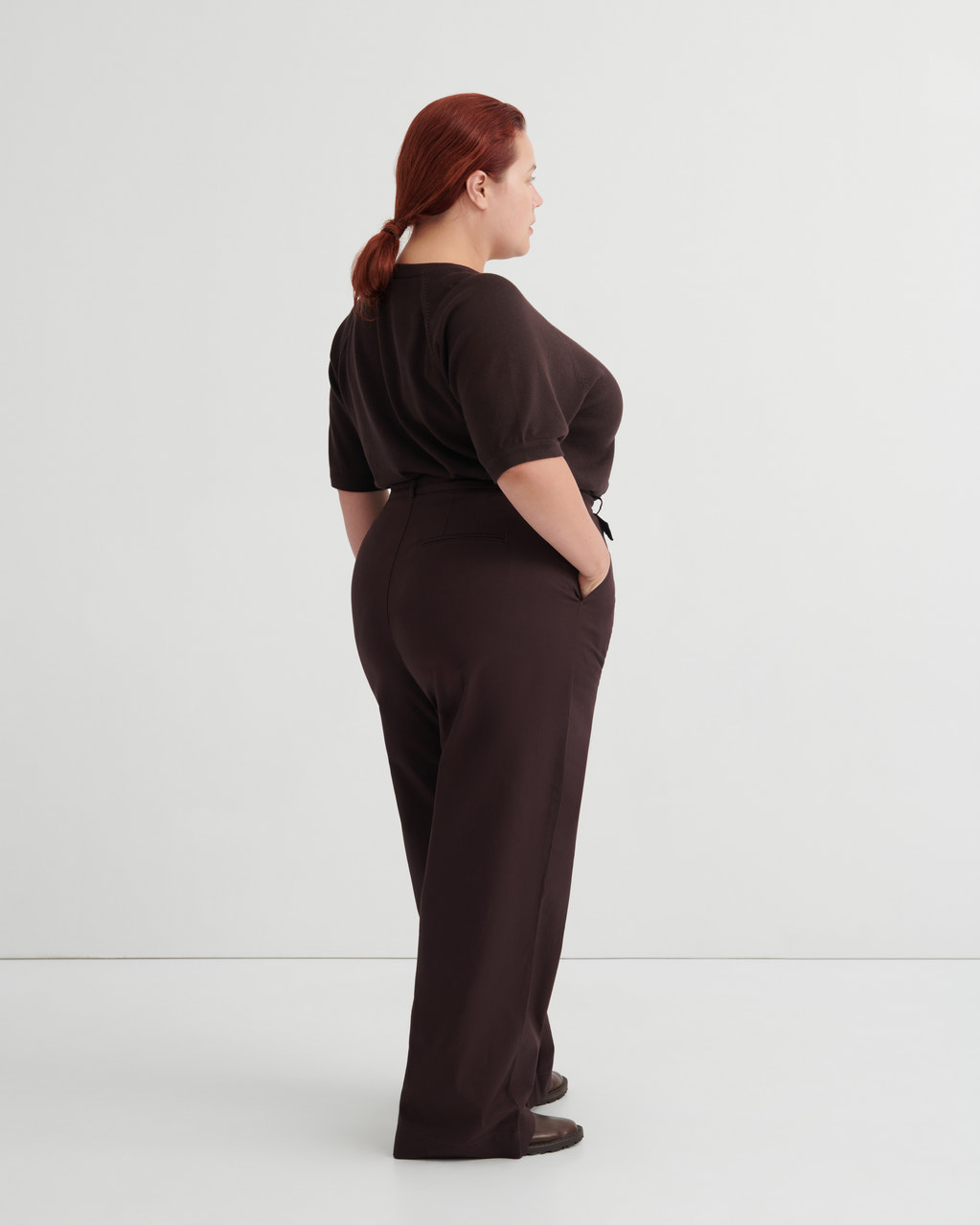 A woman with red hair tied back is shown from the back wearing a dark brown short-sleeve top and matching dark brown wide-leg trousers.