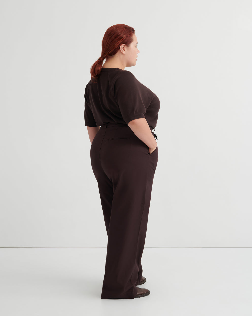 A woman with red hair tied back is shown from the back wearing a dark brown short-sleeve top and matching dark brown wide-leg trousers.