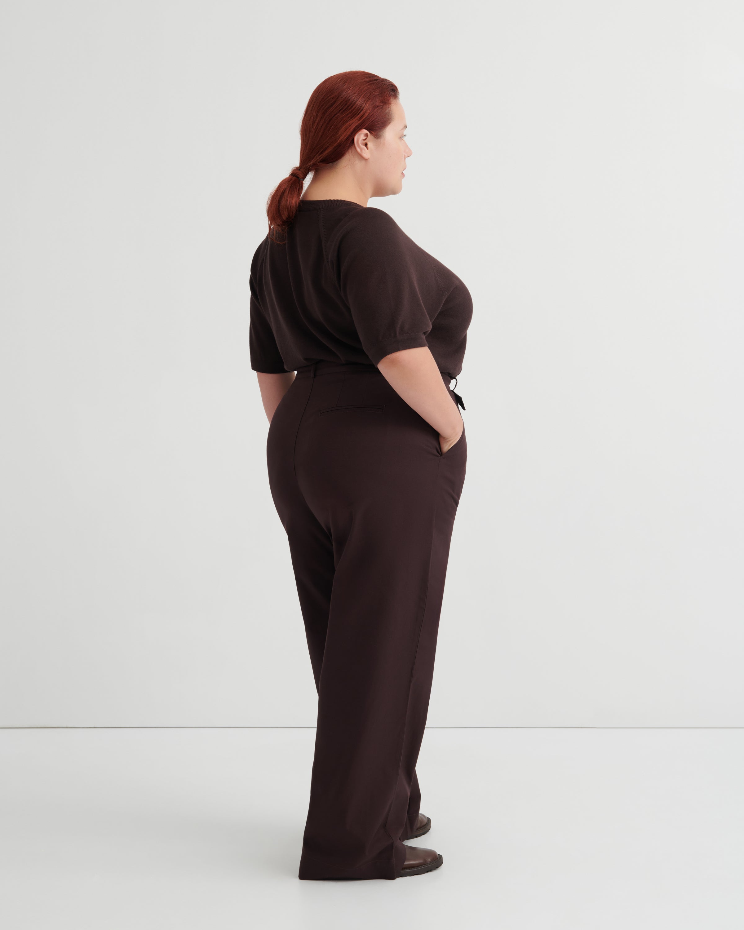 A woman with red hair tied back is shown from the back wearing a dark brown short-sleeve top and matching dark brown wide-leg trousers.
