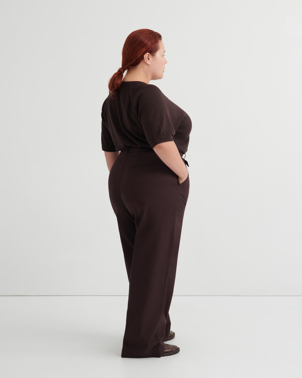 A woman with red hair tied back is shown from the back wearing a dark brown short-sleeve top and matching dark brown wide-leg trousers.