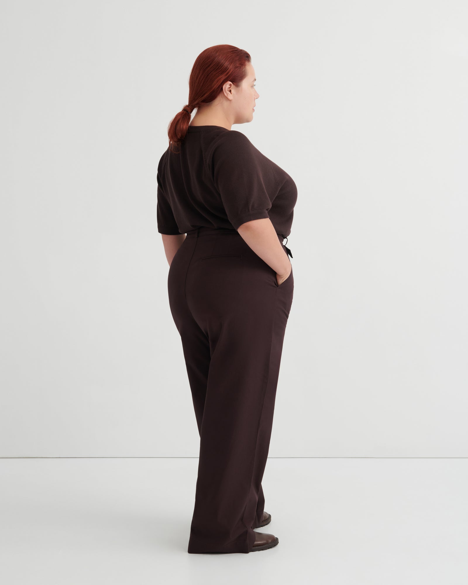 A woman with red hair tied back is shown from the back wearing a dark brown short-sleeve top and matching dark brown wide-leg trousers.