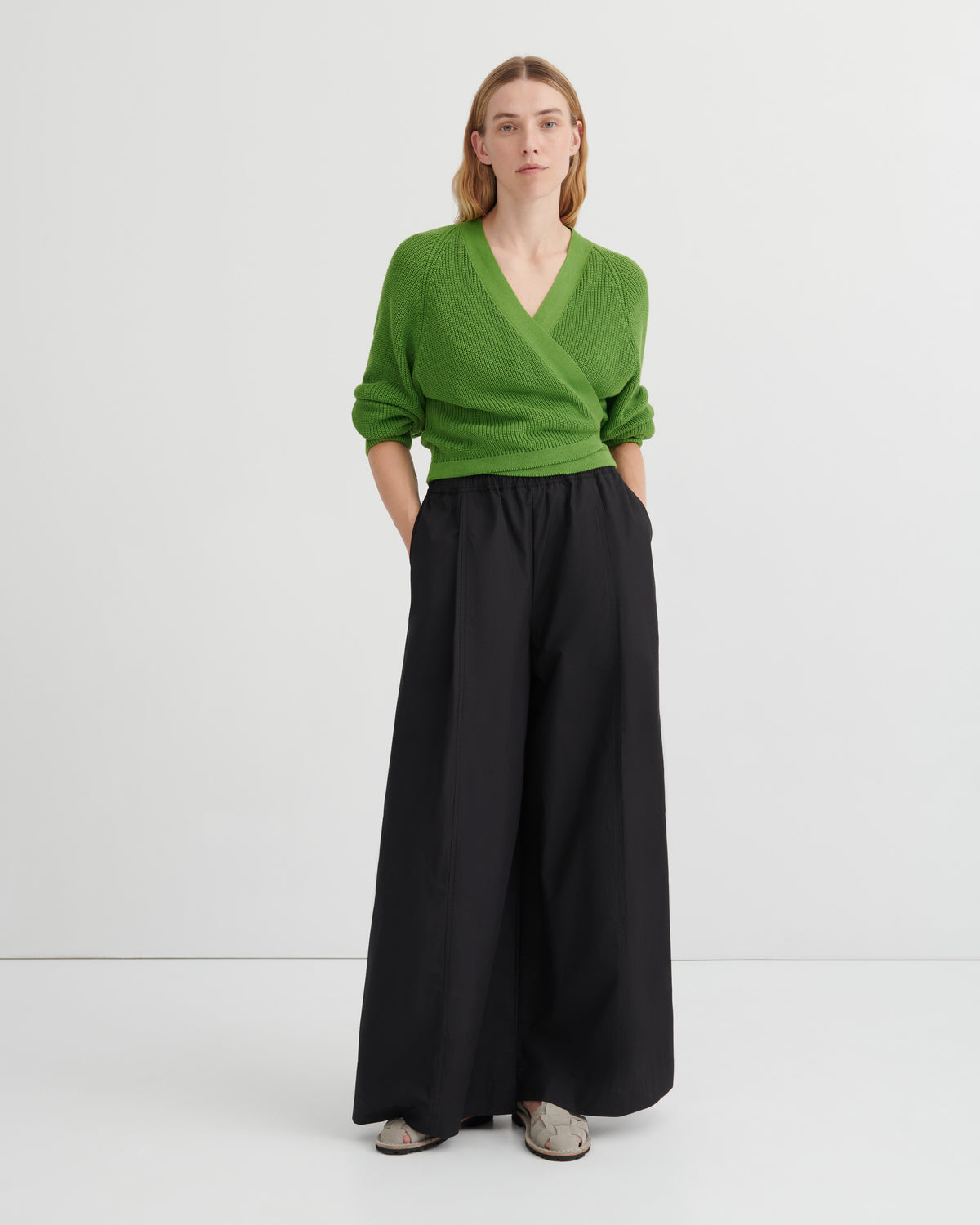 A woman stands against a plain light gray studio background wearing a bright green wrap-style V-neck sweater and wide-leg black trousers, with gray slip-on shoes.