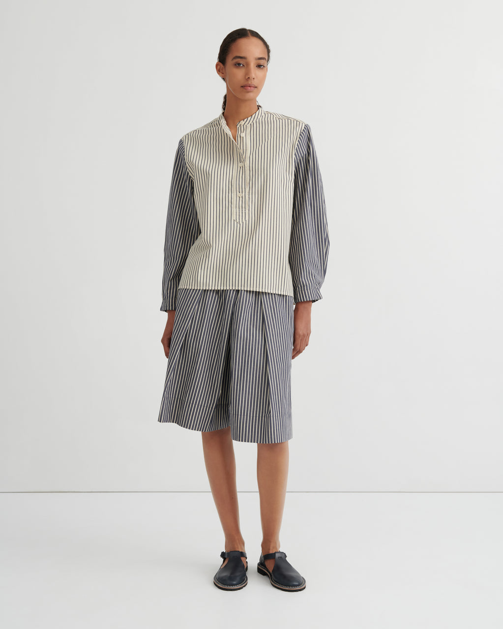 A model stands against a plain white studio background wearing a matching striped two-piece set consisting of a button up shirt and pleated shorts. The shirt has a cream and white vertical stripe pattern on the front and solid dark gray sleeves, while the shorts are entirely dark gray with thin white vertical stripes.