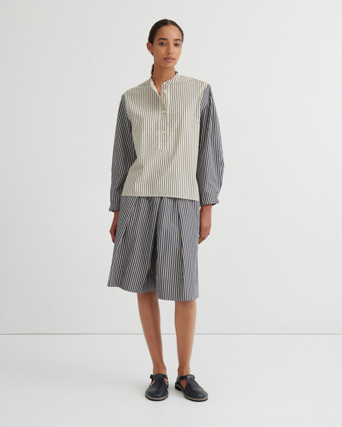 A model stands against a plain white studio background wearing a matching striped two-piece set consisting of a button up shirt and pleated shorts. The shirt has a cream and white vertical stripe pattern on the front and solid dark gray sleeves, while the shorts are entirely dark gray with thin white vertical stripes.